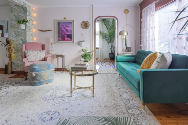 Una sala de estar acogedora con un sofá azul verdoso, un sillón rosa y un reposapiés azul. Hay una mesa de centro de vidrio y un espejo en la pared.