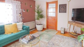 Ein Wohnzimmer mit einem türkisblauen Sofa, einem Couchtisch mit Büchern, einer großen Topfpflanze, einer Holztür und einem Fernseher auf einem Holzboden.