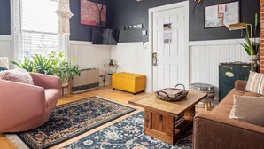 A cozy living room with a pink sofa, a yellow ottoman, and a wooden coffee table. There is a large window with blinds, a hanging light fixture, and a black and white wall with a framed picture.