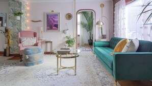A cozy living room with a teal sofa, a pink armchair, and a blue ottoman. There is a glass coffee table and a mirror on the wall.