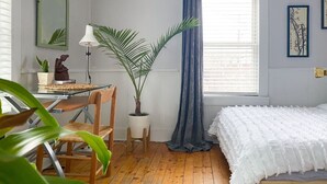 A bedroom with a wooden floor, a bed with white bedding, a desk with a lamp, a chair, a potted plant, and a window with blue curtains.