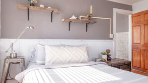 A bedroom with a bed, a nightstand, a lamp, a wooden door, and two wall-mounted shelves with books and a vase. - Bohemian Penthouse | 1min Walk Restaurants + Shops (Winooski)
