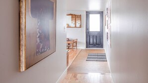 A hallway with a wooden floor, a framed picture on the left, a door at the end, and a chair in the middle.