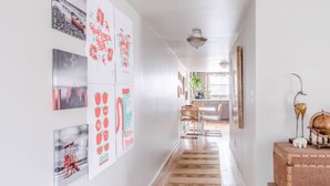 A hallway with artwork on the walls, a wooden floor, and a kitchen area in the distance.