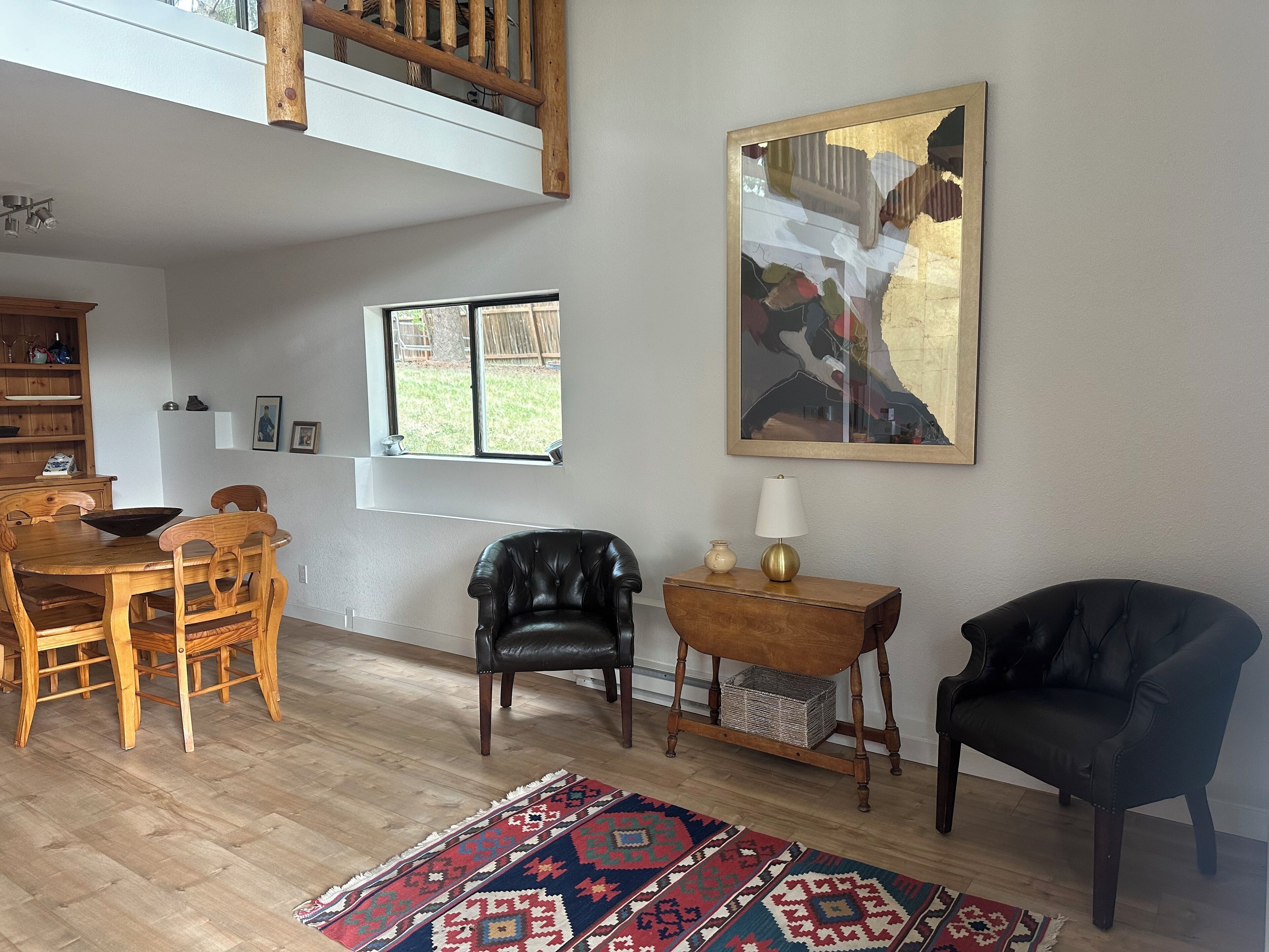 Un vaste espace de vie comprenant une table à manger en bois, deux fauteuils en cuir, un fauteuil en cuir noir, une petite table d’appoint en bois avec une lampe, et un tapis aux couleurs vives.