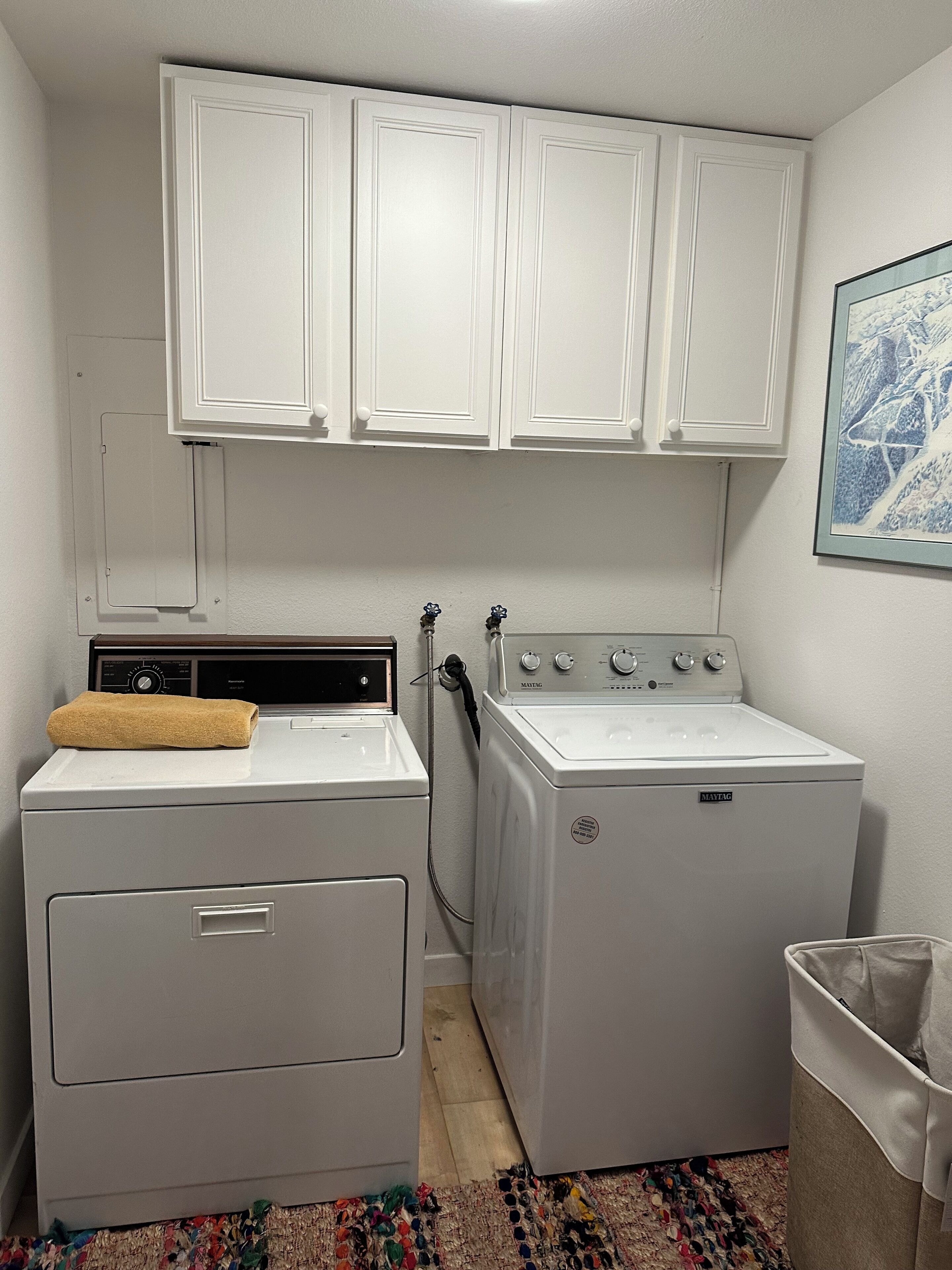 A laundry room with a front-loading washer and dryer, white cabinets above, and a small trash bin to the right.