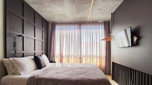 A modern bedroom with a large bed, a headboard with built-in lighting, a bedside table, a wall-mounted TV, and a window with sheer curtains. - Setai Yacht (João Pessoa)