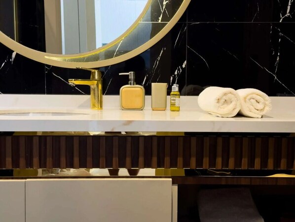 A bathroom vanity with a gold faucet, a round mirror, and toiletries including a soap dispenser, a lotion bottle, and two rolled towels.