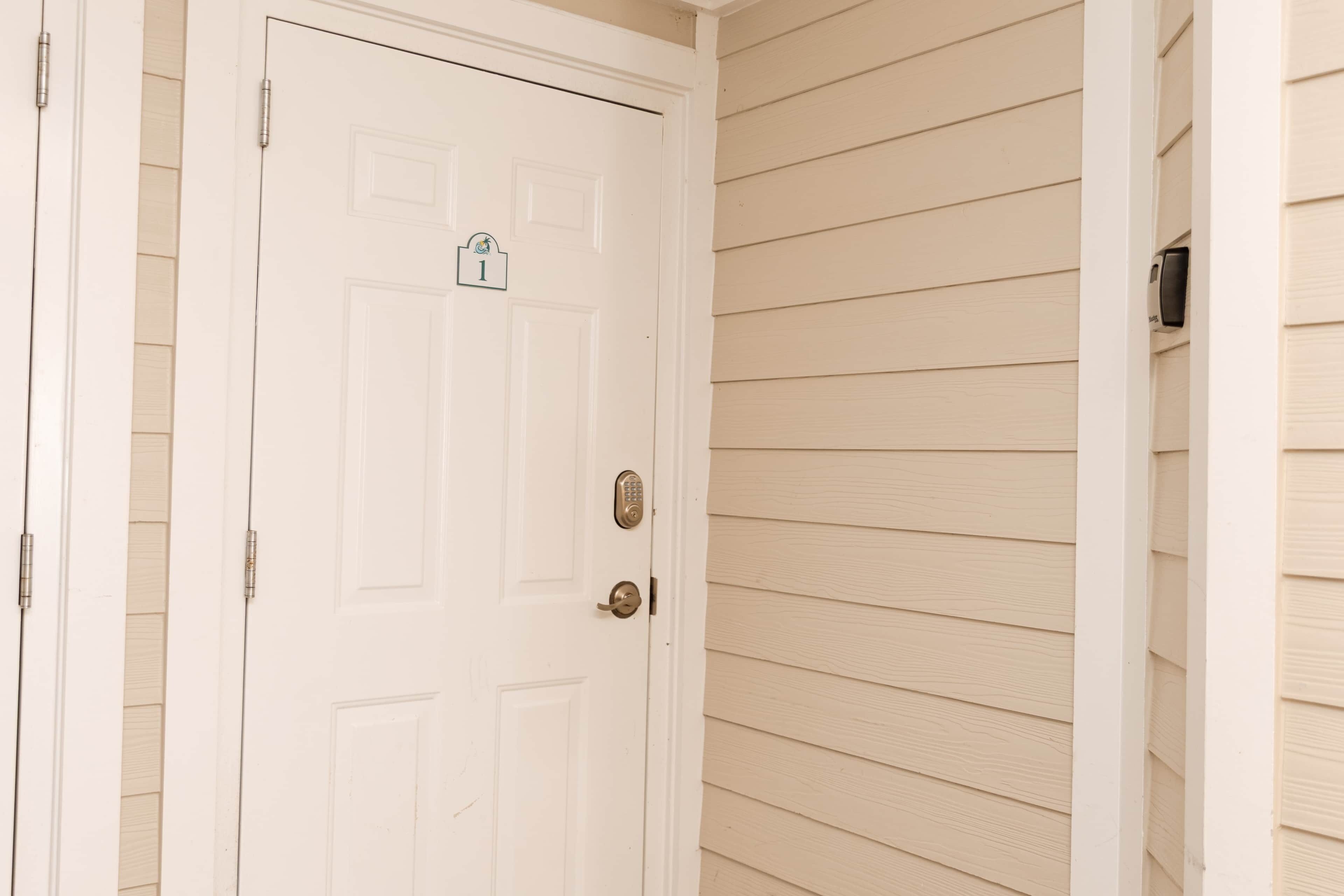 Una porta blanca amb un pany i una placa amb un número, situada en una paret exterior beix.