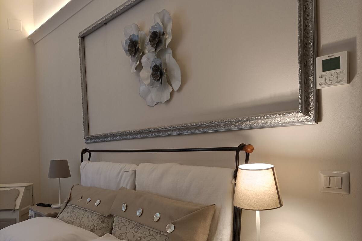 A neatly arranged bedroom with a bed, bedside tables, a lamp, and a wall-mounted thermostat.