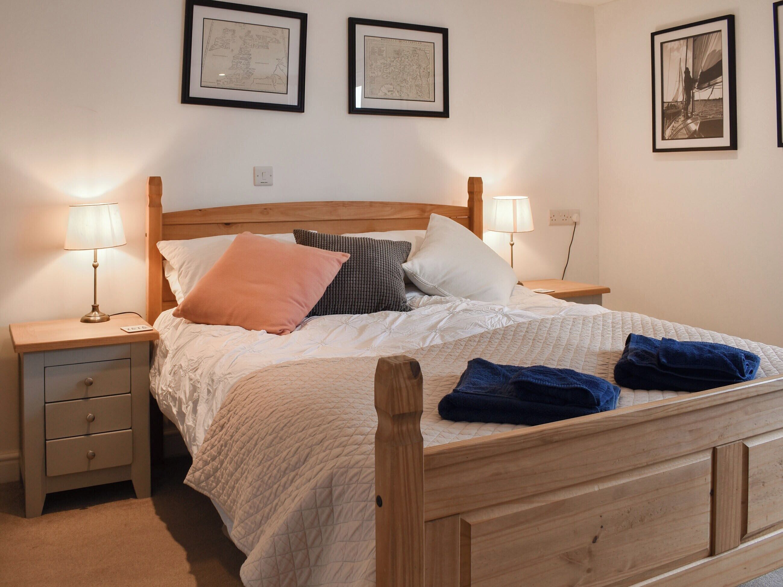 A neatly made bed with a quilted blanket, two bedside lamps, a wooden headboard, and two framed maps on the wall.