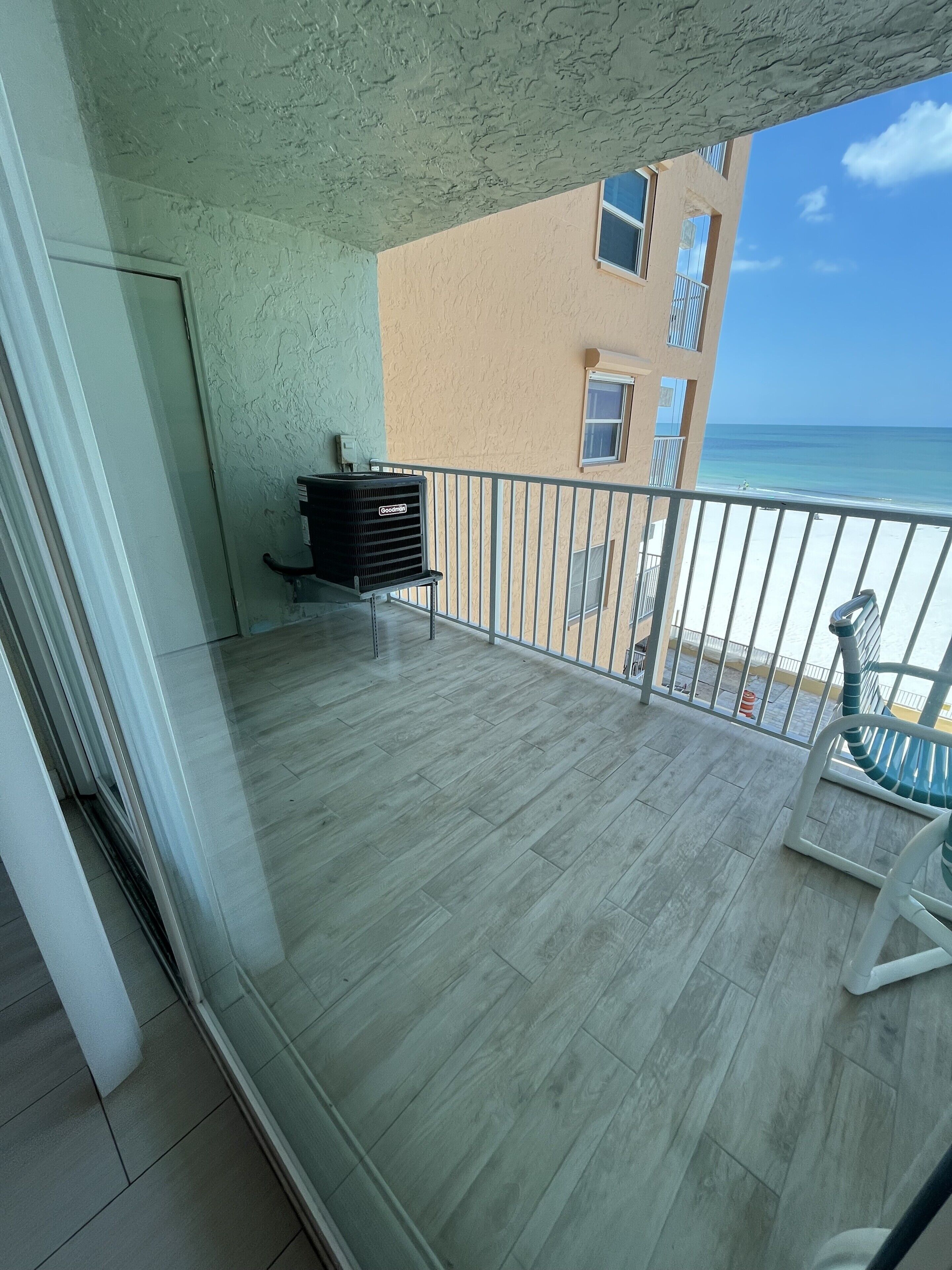 A balcony with a view of the beach, a black air conditioning unit, and a white chair.