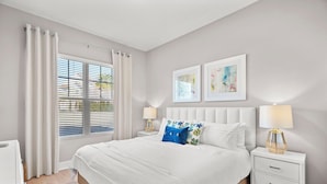 Une chambre avec un grand lit, des rideaux blancs, une table de chevet avec une lampe et deux tableaux encadrés accrochés au mur.