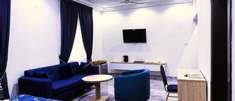 A modern hotel room with a blue sofa, a round coffee table, a bed with a star-patterned duvet, a desk with a flat-screen TV, and a chair.