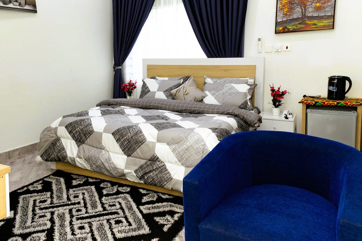 A bedroom with a bed, a blue chair, a nightstand, a coffee machine, and a painting on the wall.