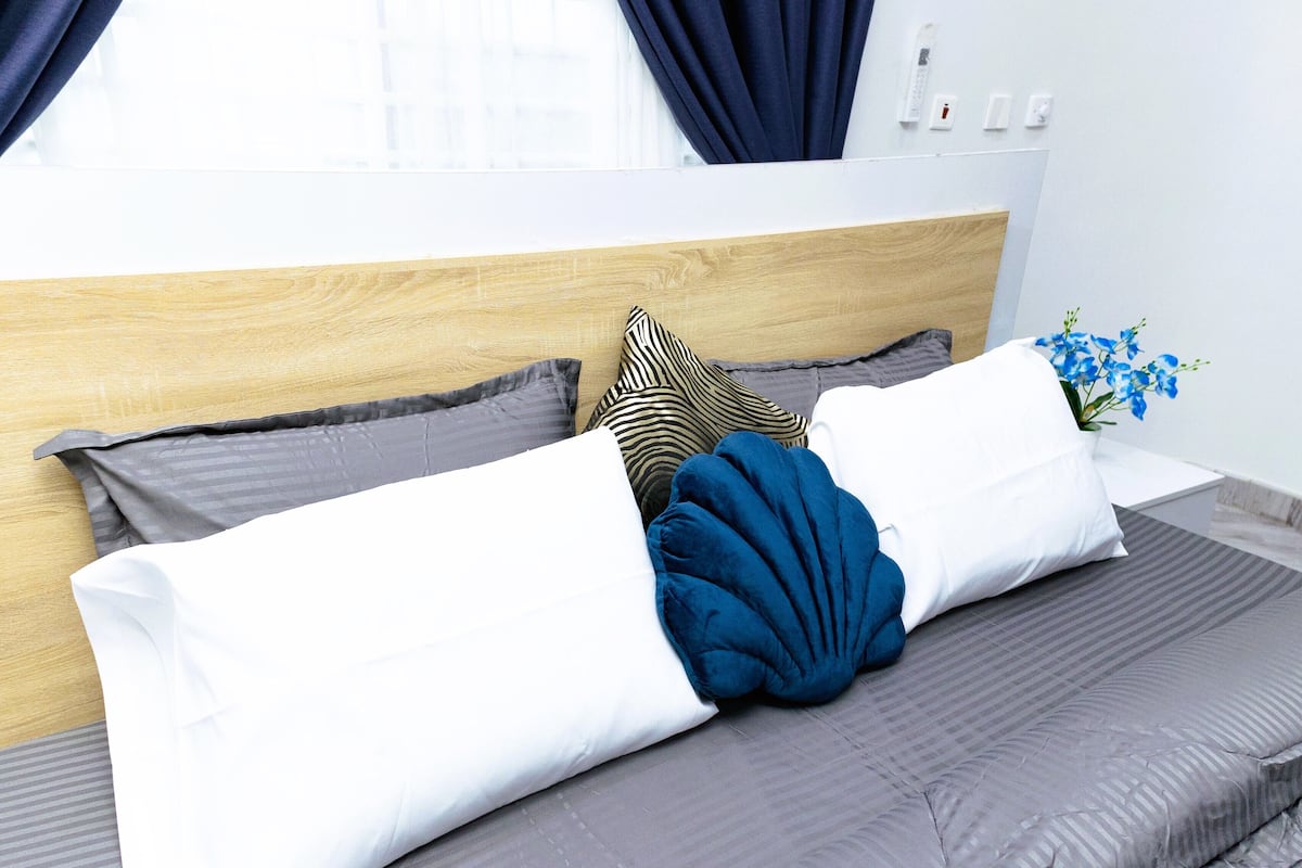 A neatly made bed with grey bedding, a wooden headboard, and a vase with blue flowers on the nightstand.