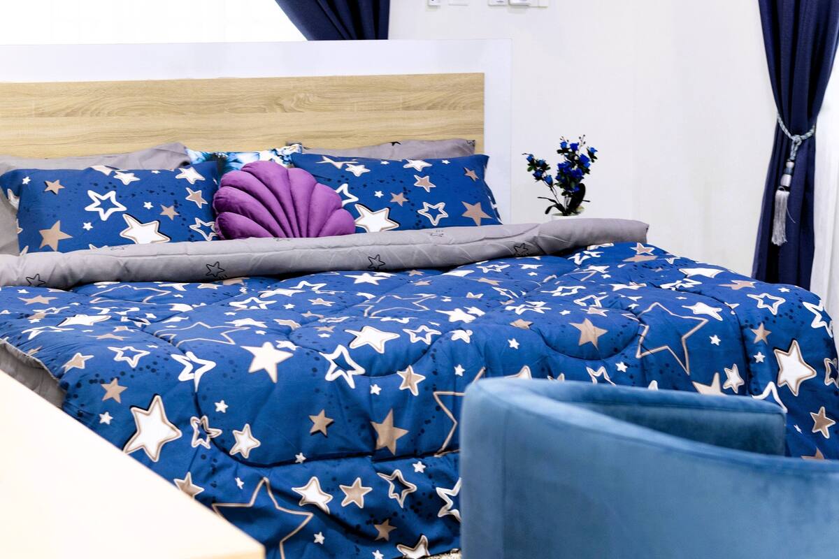 A bedroom with a bed covered in a blue star-patterned duvet, a wooden headboard, and a vase with flowers on the nightstand.