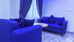 A modern living room with a blue sofa set, a round coffee table, and a large window with curtains. - Firnic Luxurious Private Hotel (Kwabenya)