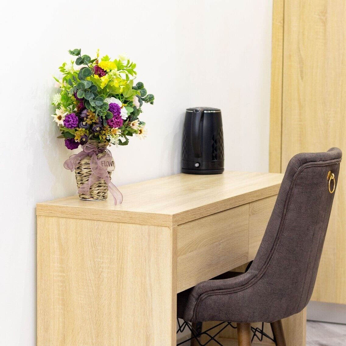 A wooden desk with a woven basket vase containing a bouquet of flowers, a black speaker, and a grey upholstered chair.