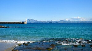 A coastal scene with clear blue water, rocky shoreline, and distant mountains under a clear sky.