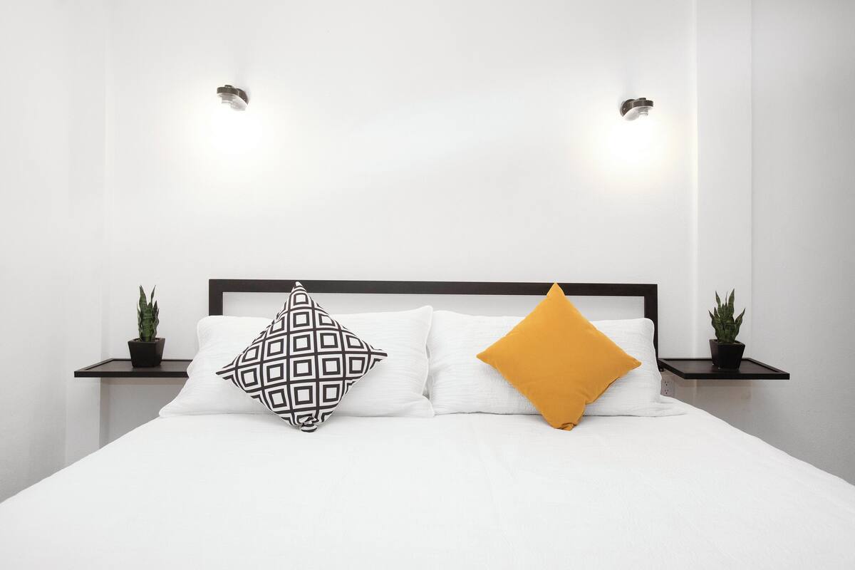 Un lit bien fait, avec une literie blanche et deux oreillers : l’un à motifs géométriques et l’autre de couleur jaune moutarde. Deux petites plantes en pot sont placées de chaque côté du lit.