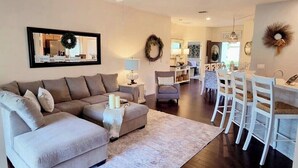 A living room with a sectional sofa, a small table, and a coffee table. There is a dining area with white chairs and a chandelier.