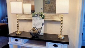 A room with a black console table, two lamps, a mirror, and a sign that reads 'BE OUR GUEST.'