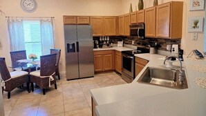 A modern kitchen with wooden cabinets, stainless steel appliances, a dining area with wicker chairs, and a clock on the wall.
