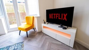 A modern living room with a large flat-screen TV mounted on a white cabinet, a yellow armchair, and a view of a fenced outdoor area through a sliding glass door. - Collingwood Lodge, Birmingham with FREE Parking (West Midlands)