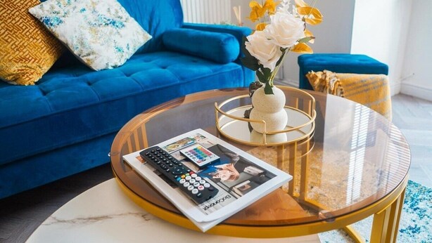 A living room with a blue sofa, a glass coffee table, a magazine, a remote control, and a vase with flowers. - Collingwood Lodge, Birmingham with FREE Parking (West Midlands)