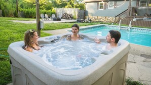 A hot tub with three people in it, surrounded by a backyard with a pool, chairs, and a house.