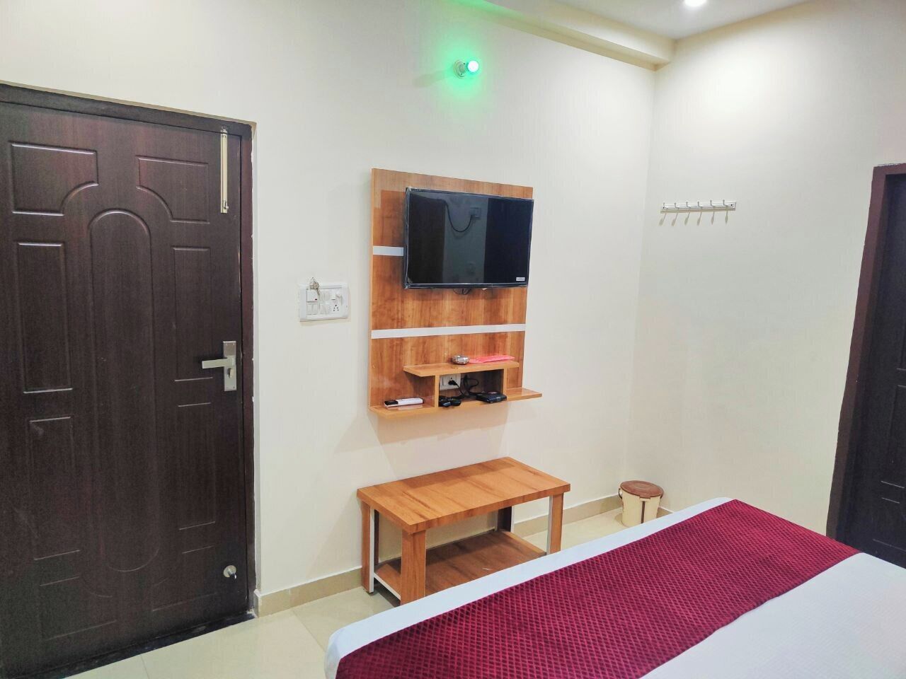 A bedroom with a wooden door, a bed with a red and white bedspread, a small wooden table, a mounted television, and a wall-mounted shelf with a few items.