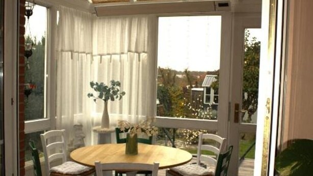 A conservatory with a round wooden table and white chairs, surrounded by white curtains and a potted plant.