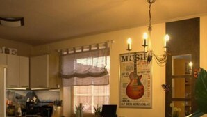 A compact kitchen with white cabinets, a wooden countertop, and a tiled floor. There is a poster on the wall and a chandelier hanging above the counter.