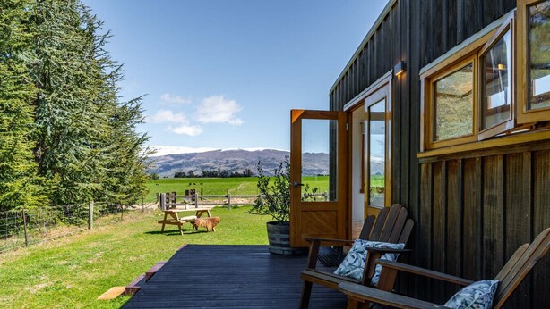 A wooden deck with two chairs, a table, and a door leading to a view of a green field and mountains.