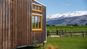 A wooden cabin with a window, set against a backdrop of snow-capped mountains and a green field.