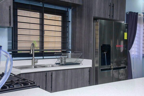 A modern kitchen with a stainless steel refrigerator, a sink with a faucet, and a window with blinds. - Montana Villa-ochorios-sleeps6-aircon-pool-tv-wifi (Ocho Rios)