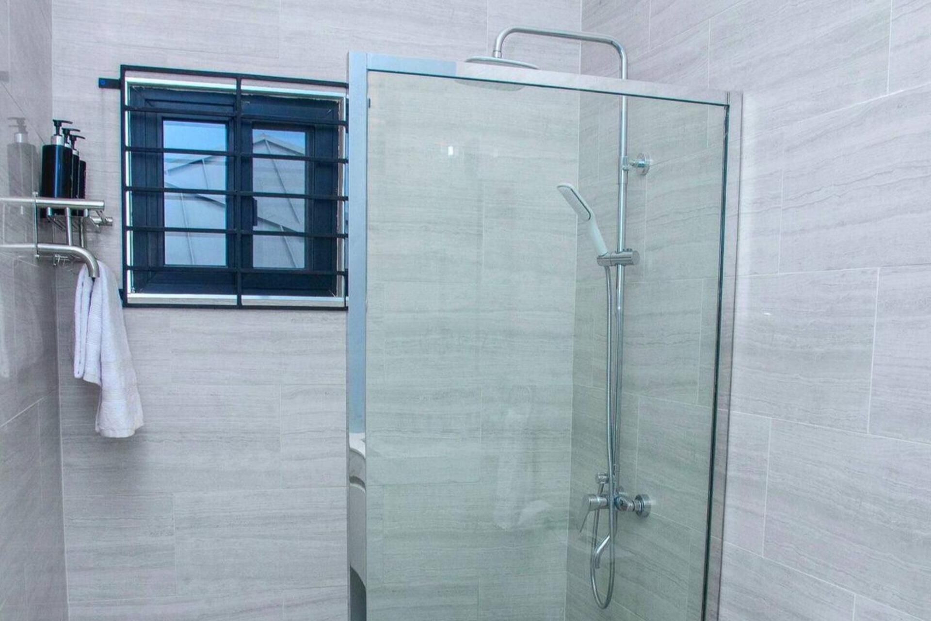 A modern bathroom with a glass-enclosed shower, a window with blinds, and a towel rack with toiletries.