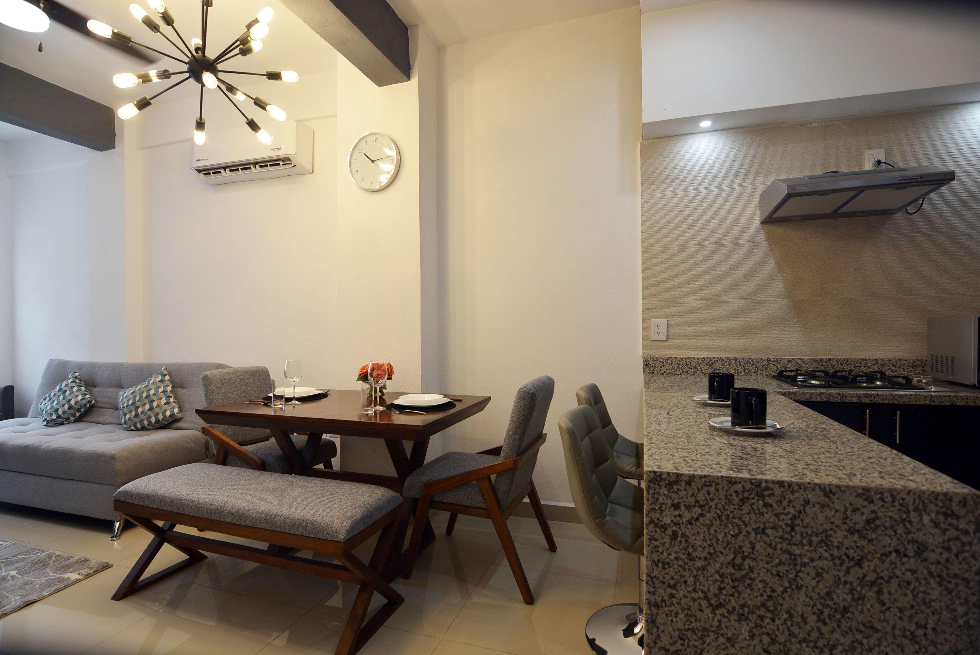 A modern living room with a dining table, grey sofa, and a kitchen area with granite countertops.