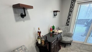 A living room with two patterned armchairs, a small coffee table, and a cabinet with bottles. There are wall-mounted shelves and a sliding glass door leading to a balcony.