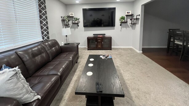 A living room with a brown leather sofa, a wooden coffee table, a flat-screen TV mounted on the wall, and a dining area with a table and chairs.