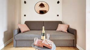 A grey sofa with two pink cushions, a bottle of champagne, a glass, and a box of chocolates on a small table in front of it. - TheBlueStay 39 – Cosy Apartment Buttes-Chaumont (Paris)