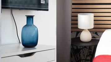 Ein modernes Zimmer mit einer blauen Vase auf einer weißen Kommode, einer weißen Lampe auf einem kleinen runden Tisch und einem an der Wand montierten Fernseher.