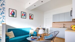 A modern living room with a blue sofa, two wicker chairs, a dining table with chairs, and a kitchen area with cabinets and a fruit bowl.