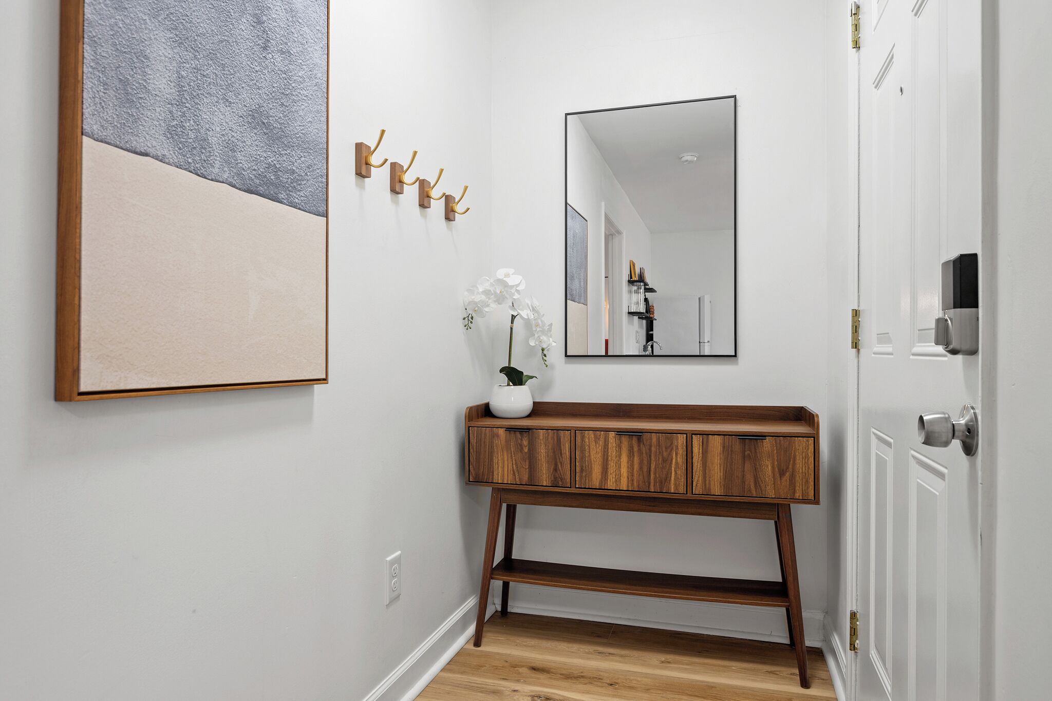 Een gang met een houten console tafel, een witte vaas met een plant en een groot abstract schilderij aan de muur.