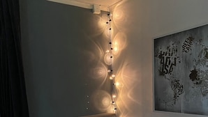 A bedroom with a bed, pillows, a headboard, a string of lights, a wall art piece, and a curtain.