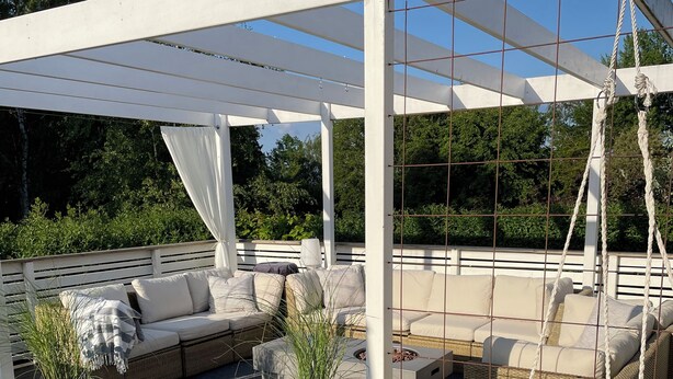A covered outdoor seating area with a wooden deck, white pergola, and hanging chairs.