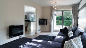 A modern living room with a large sofa, a flat-screen TV mounted on the wall, and a view of a garden through a sliding glass door. - Superb 4BD Stay in Wyton and Houghton Village (Huntingdon)