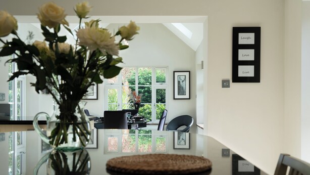 A dining area with a glass table, chairs, and a vase of flowers. There are framed pictures on the wall and a view of greenery outside. - Superb 4BD Stay in Wyton and Houghton Village (Huntingdon)