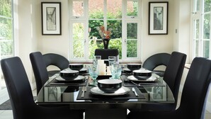 A dining table with black chairs, set with plates, bowls, glasses, and a candle. There are framed pictures on the walls and a window with a view of greenery. - Superb 4BD Stay in Wyton and Houghton Village (Huntingdon)